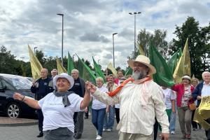 Senioriada. Korowód seniorów przechodzi przez parking przed pływalnią Wodny Raj. W pierwszej parze idzie radna Rady Miejskiej Seniorów i wodzirej.