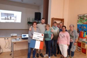 Wnętrze Centrum Aktywności Seniora. Wolontariusz europejski stoi w otoczeniu uśmiechniętych seniorek. Jedna z nich trzyma flagę Francji i transparent.