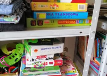 Stalowy regał z kolorowymi pudełkami gier planszowych dla dzieci i dorosłych, m.in. Słowny Ekspres, Krzyżówka Liter, Gra planszowa, Asteriks, Love Puzzle. Obok gier widoczne złożone ubrania.