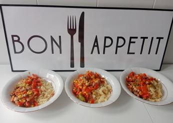 Na zdjęciu widać trzy białe głębokie talerze, w których jest makaron świderki, boczek i warzywa. Talerze stoją na tle tablicy z napisem Bon Apetit.