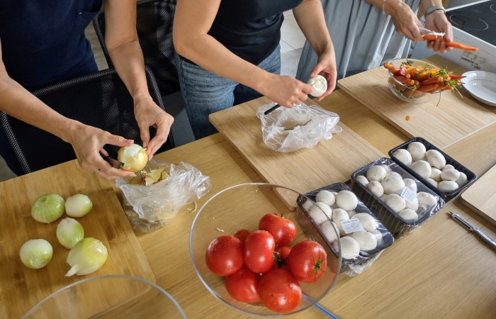 Osoby obierają cebulę, pieczarki i marchewki na drewnianych deskach. Na stole leżą miski z pomidorami i opakowania pieczarek.