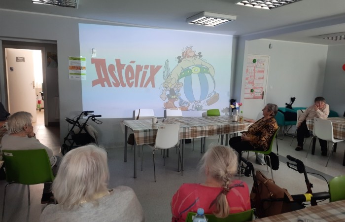 Wnętrze Dziennego Domu Senior Wigor. Widać seniorów patrzących w kierunku ściany. Na ścianie wyświetlany jest slajd z prezentacji o komiksie Asterix przygotowanej przez Żuliet Żama - francuską wolontariuszkę Europejskiego Korpusu Solidarności.