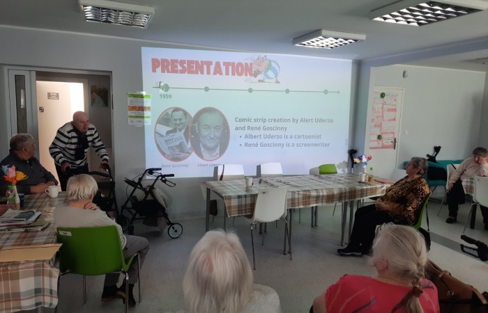 Wnętrze głównej sali Dziennego Domu Senior Wigor. Widać seniorów siedzących przy stolikach zwróconych w kierunku ściany. Przez otwarte drzwi do sali wchodzi o balkoniku jeden z seniorów. Na ścianie wyświetlany jest kolejny slajd z prezentacji Żuliet.