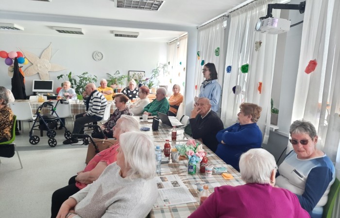 Zdjęcie przedstawia widok głównej sali w Dziennym Domu Senior Wigor oświetlonej słońcem. Seniorzy siedzą przy dwóch długich stołach ustawionych do siebie prostopadle. Pomiędzy siedzącymi i oglądającymi seniorami widać stojącą i opowiadającą Żuliet.