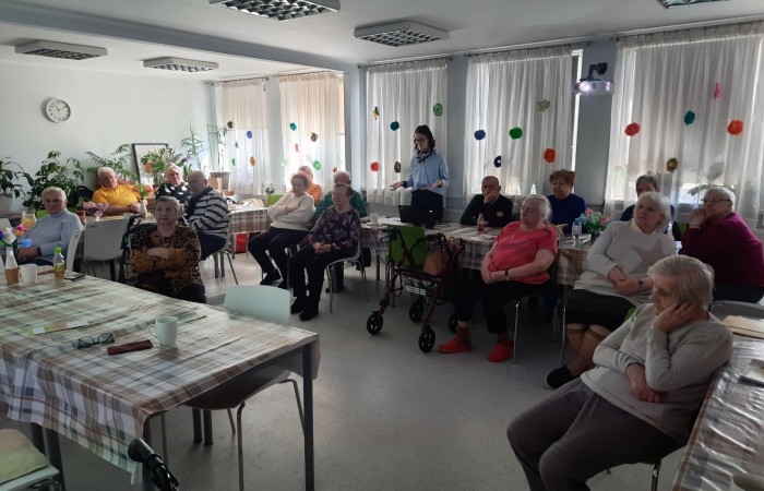 Wnętrze Dziennego Domu Senior Wigor. Zdjęcie przedstawia widok głównej sali. Okna są zasłonięte roletami. Szesnaścioro seniorów siedzących przy stołach patrzy w kierunku ściany, na której wyświetlane są slajdy z prezentacji Żuliet Żama.