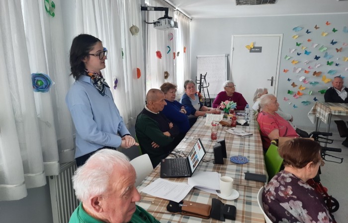 Wnętrze Dziennego Domu Senior Wigor. Zbliżenie na Żuliet Żama - wolontariuszkę Europejskiego Korpusu Solidarności, która pokazuje swoją prezentację o Asteriksie. Żuliet stoi przy stole, na którym ustawiony jest laptop. Obok niego leżą notatki Żuliet.