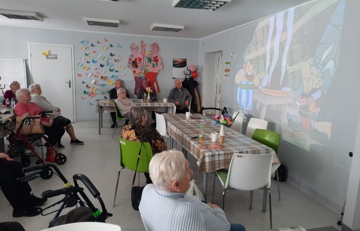 Wnętrze Dziennego Domu Senior Wigor. Żuliet Żama prezentuje kolejny slajd swojej prezentacji, na którym widać Obeliksa i Asteriksa stojących przy drewnianym stole, na którym paruje świeżo upieczone mięso świni. Obaj Galowie jedzą pieczone udo.