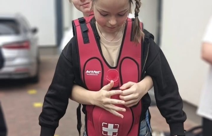 Ostatni piąty punkt gry miejskiej, czyli podwórko przed Klubem Młodzieży Nowy Świat dziesięć. Na pierwszym planie widać dwie dziewczynki wykonujące zadanie. Jedna z nich ma na sobie specjalną kamizelkę, a druga stojąca za nią obejmuje ją rękami.