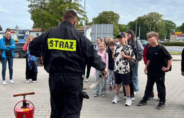 Zdjęcie przedstawia punkt startowy gry miejskiej pod tytułem Ratujemy bo umiemy. Na pierwszym planie widać strażaka tłumaczącego grupie uczniów konkurencję, do której za chwilę przystąpią. Obok strażaka stoi czerwona hydronetka wodna z żółtą etykietą.