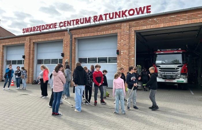 Na zdjęciu widać plac przed Swarzędzkim Centrum Ratunkowym. Grupa kilkunastu uczniów i uczennic zapoznaje się z wężem strażackim, którym za chwilę będzie wykonywać zadanie. W tle widać otwarty garaż remizy, a w nim przód dużego samochodu strażackiego.