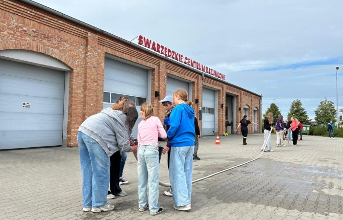 Na tle Swarzędzkiego Centrum Ratunkowego zespół składający się w większej części z dziewczynek wykonuje zadanie polegające na wlewaniu wody z wiadra do węża i gaszeniu udawanego pożaru.