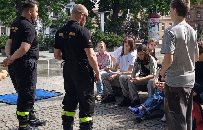 Trzeci punkt gry miejskiej na placu Niezłomnych. Dwóch strażaków tłumaczy drużynie siedzącej na ławce kolejną konkurencję. Obok strażaków stoi jeden uczeń. W tle widać tryskające fontanny, słup ogłoszeniowy, wysokie zielone drzewa i stare kamienice.
