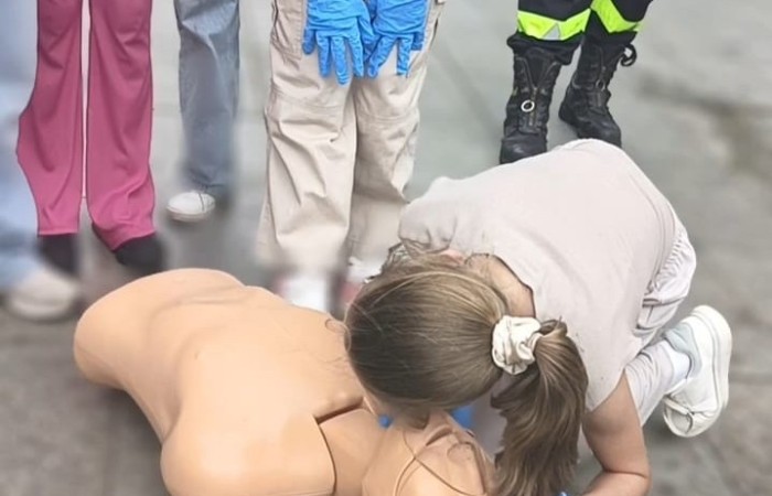 Płyta placu Niezłomnych. Jedna z zawodniczek klęcząc odchyla głowę fantomowi i sprawdza oddech. Nad nią stoją inne zawodniczki i strażak. Wszystkie osoby wykonujące ćwiczenie mają na rękach niebieskie rękawiczki.
