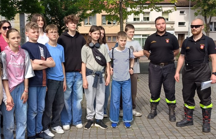 Zdjęcie grupowe wykonane na placu Niezłomnych na trzecim punkcie gry miejskiej pod tytułem Ratujemy bo umiemy. Zdjęcie przedstawia dziewięcioro zawodników, nauczycielkę i dwóch strażaków. Za ich plecami widać zielone drzewa i stare kamienice.