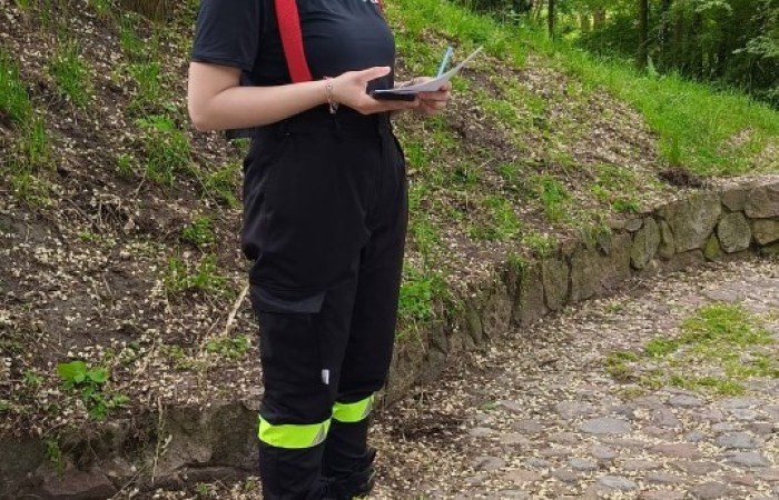 Czwarty punkt gry miejskiej, czyli Boisko pod Kasztanami przy ulicy świętego Marcina. Na chodniku z otoczaków na tle wzgórza porośniętego zielonymi drzewami i krzewami stoi strażaczka, która w ręku trzyma kartki, telefon i długopis.