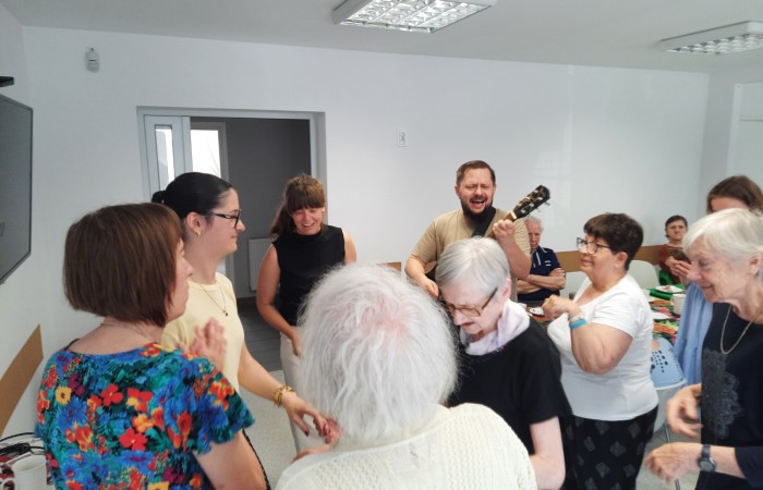 Pożegnanie Żuljet Żama w Dziennym Domu Pomocy. Pracownik Domu Rafał Wójtowicz gra na gitarze, a seniorki, członkini kadry Patrycja Górecka i wolontariuszka Żuljet tańczą w kółku.