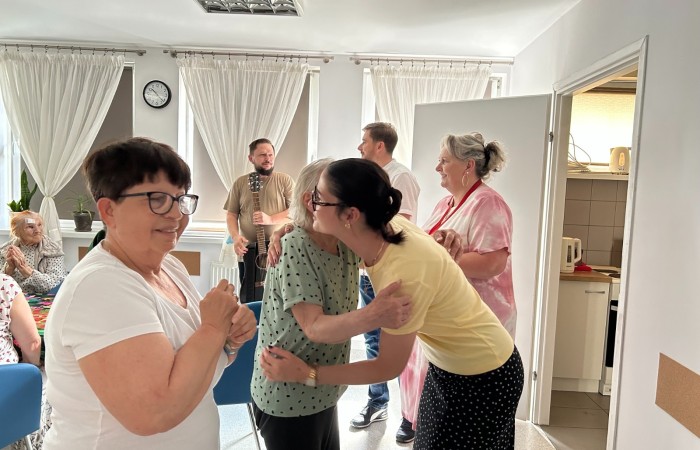 Wnętrze Dziennego Domu Pomocy. Pożegnanie Żuljet Żama wolontariuszki Europejskiego Korpusu Solidarności. Wolontariuszka przytula się do seniorki składającej jej życzenia. Druga seniorka czeka w kolejce do Żuljet. W tle widać troje pracowników.