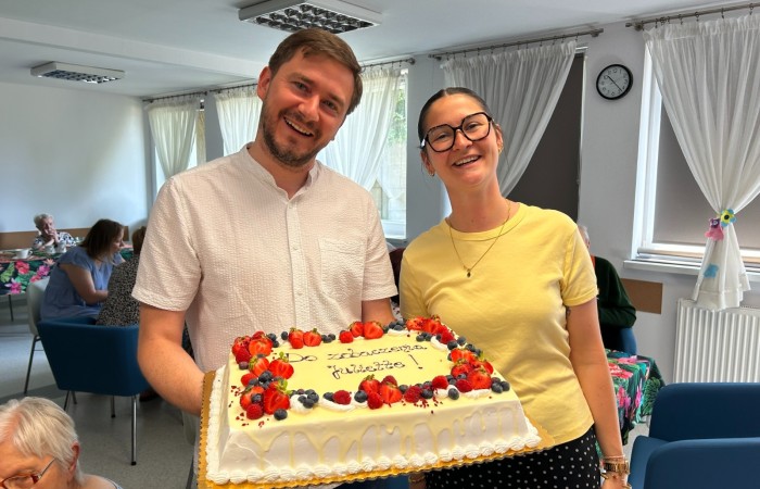 Wolontariuszka Żuljet Żama i koordynator projektu Dariusz Bugajski patrzą uśmiechnięci prosto w obiektyw trzymając w rękach duży uroczysty tort z napisem: Do zobaczenia Żuljet. W tle widać główną salę Dzienneo Domu Pomocy.