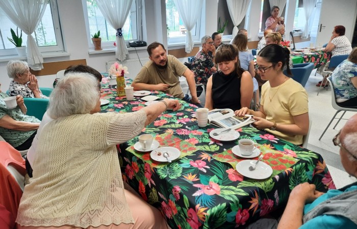 Pracownik Dziennego Domu Pomocy Rafał Wójtowicz, członkini kadry Patrycja Górecka oraz wolontariuszka Żuljet siedzą przy stole z seniorami i oglądają album, który Żuljet dostała w prezencie.