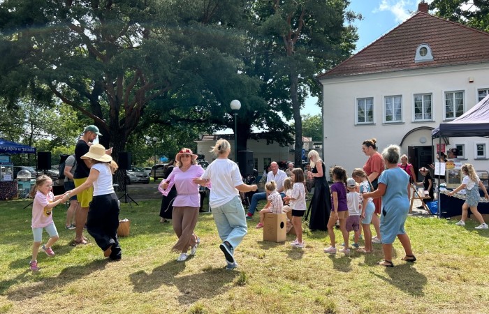 Zdjęcie wykonano na trawie przy budynku Ośrodka Kultury w Swarzędzu. Na pierwszym planie widoczne radośnie tańczące kobiety z dziećmi. W tle orkiestra flażoletowa, namioty z przekąskami i wysokie drzewa.