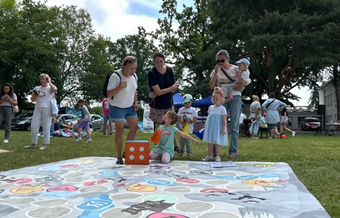 Zdjęcie wykonano na trawie przy budynku Ośrodka Kultury w Swarzędzu. Na pierwszym planie gra wielkoformatowa z dużą kostką do gry i pionkami. Nad grą pochylają się trzy kobiety, jedna z dzieckiem na rękach oraz troje dzieci.