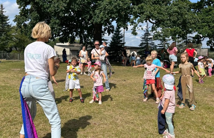 Zdjęcie wykonano na trawie wśród wysokich liściastych drzew. Na pierwszym planie widoczne radośnie tańczące dzieci oraz pani instruktor odwrócona tyłem do obiektywu.