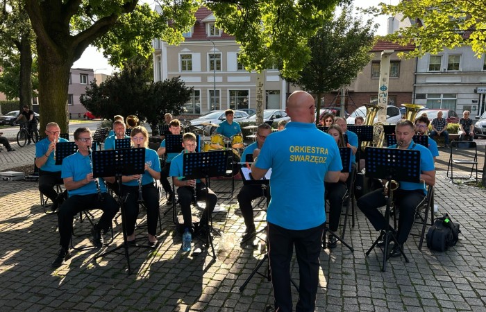 Kilkanaścioro muzyków ze Swarzędzkiej Orkiestry Dętej pod batutą stojącego tyłem do obiektywu Łukasza Gowarzewskiego koncertują na Placu Niezłomnych w otoczeniu liściastych drzew i kamiennic.