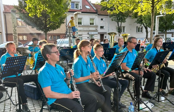 Kilkanaścioro muzyków ze Swarzędzkiej Orkiestry Dętej koncertują na Placu Niezłomnych w otoczeniu liściastych drzew i kamiennic. Na pierwszym planie muzycy grający na fletach prostych.