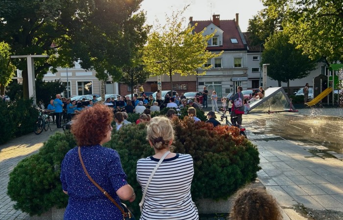 Koncert Swarzędzkiej Orkiestry Dętej na Plantach. Na pierwszym planie odwrócone tyłem trzy kobiety, a w głębi zdjęcia widać muzyków, zebranych ludzi i bawiące się dzieci.
