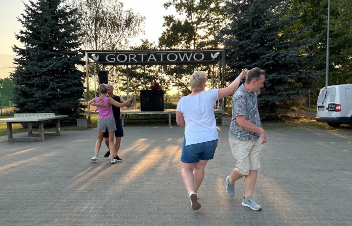 Potańcówka w Gortatowie. Dwie pary tańczą na placu przy świetlicy wiejskiej. W tle widać scenę, duży napis Gortatowo, drzewa i światło zachodzącego słońca.