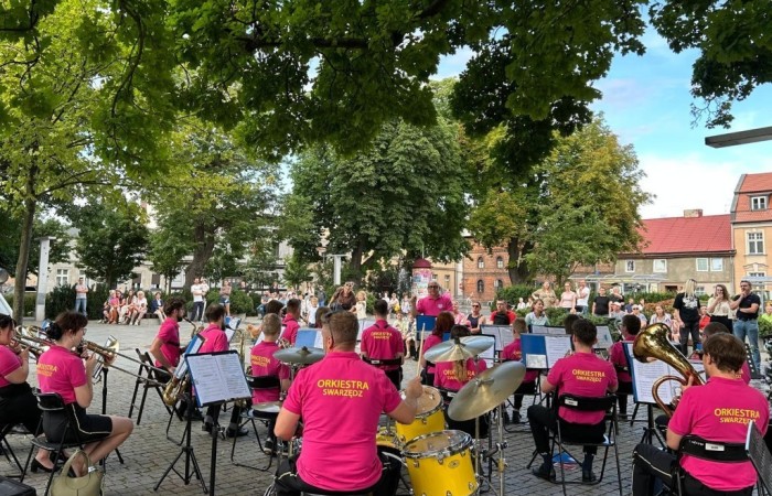 Koncert Orkiestry Dętej na Placu Niezłomnych. Na pierwszym planie widać orkiestrantów siedzących tyłem do obiektywu. W tle widać zielone drzewa, kamienice i błękitne niebo.