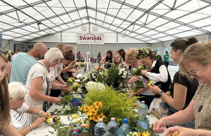 Piknik Trzech Pokoleń. Wnętrze namiotu przed sceną nad jeziorem. Przy długim stole, na którym leżą kwiaty, rośliny i ozdoby stoi około dwanaścioro osób, które plotą wianki. W tle widać baner Swarzędz dobrze trafiłeś.