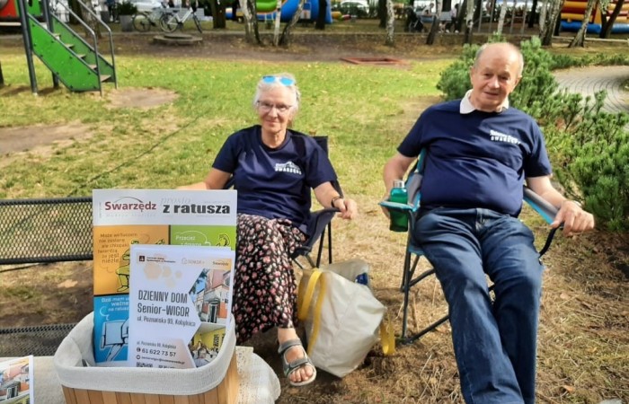Park w Kobylnicy. Na pierwszym planie widać koszyk z gazetą Prosto z Ratusza oraz ulotkami informacyjnymi na temat Dziennego Domu Senior Wigor. W centrum zdjęcia widać dwoje seniorów siedzących wygodnie na krzesełkach turystycznych. Dalej plac zabaw.