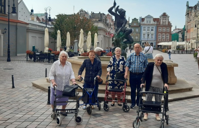 Grupa czterech seniorek z balkonikami i dwóch seniorów bez balkoników stojących przed jedną z fontann na Starym Rynku w Poznaniu. W tle widać budynek Odwachu, ogródki piwne oraz zabytkowe kamienice Starego Rynku.