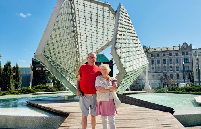 Para seniorów stoi na drewnianym pomoście przed współczesną fontanną na Placu Wolności. W tle widać zabytkowe kamienice placu. Górną połowę zdjęcia stanowi błękitne bezchmurne niebo.