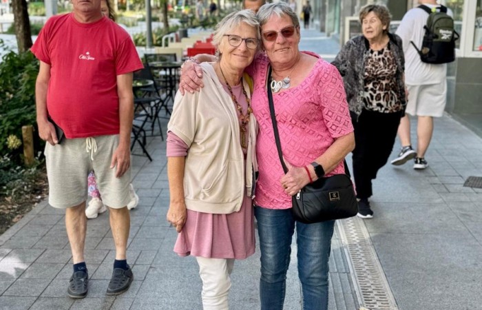 Zdjęcie przedstawia chodnik przy Placu Wolności w Poznaniu. Na pierwszym planie widać dwie przytulające się do siebie seniorki, za nimi widać dwoje innych idących seniorów. Tło stanowi szpaler zielonych drzew, ściany kamienic oraz błękitne niebo.