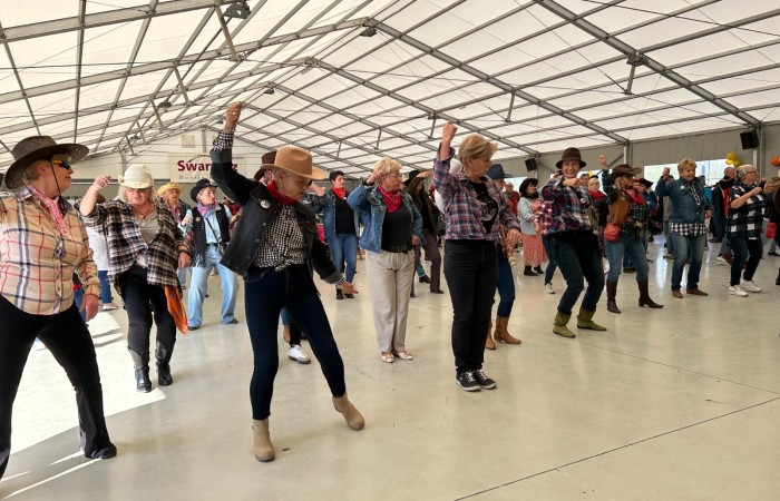 Zdjęcie przedstawia ubranych w kowbojskie stroje seniorów wspólnie tańczących. Scena rozgrywa się pod namiotem przy scenie nad jeziorem w Swarzędzu.