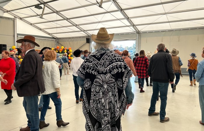 Zdjęcie przedstawia ubranych w kowbojskie stroje seniorów wspólnie tańczących. Scena rozgrywa się pod namiotem przy scenie nad jeziorem w Swarzędzu. Na pierwszym planie tyłem stoi Pan z siwymi włosami w kapeluszu kowbojskim i ponczo.