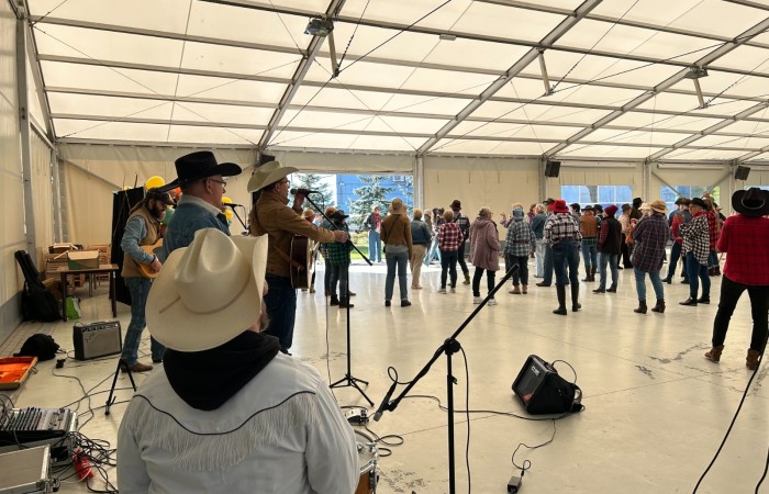 Zdjęcie przedstawia ubranych w kowbojskie stroje seniorów wspólnie tańczących. Scena rozgrywa się pod namiotem przy scenie nad jeziorem w Swarzędzu. Na pierwszym planie zespół muzyczny grający muzykę country.