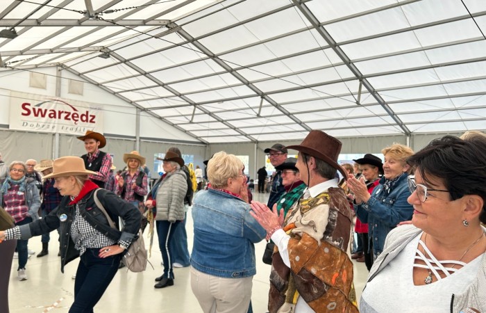 Zdjęcie przedstawia roześmianych ludzi ubranych w kapelusze, poncza, kamizelki i chustki świadczące o tym,  że tańczą na imprezie w stylu country. Impreza odbywa się pod namiotem w Swarzędzu.