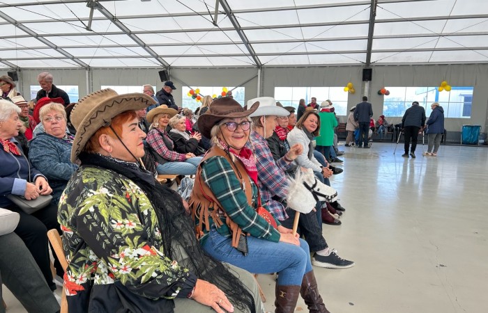 Zdjęcie przedstawia roześmianych ludzi ubranych w kapelusze, poncza, kamizelki i chustki świadczące o tym,  że uczestniczą w imprezie w stylu country. Zebrani siedzą na drewnianych krzesłach ustawionych w rzędach. Impreza odbywa się pod namiotem w Swarzędzu.