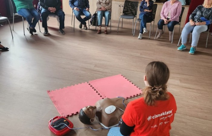 Wnętrze głównej sali w Centrum Aktywności Seniora. Na podłodze klęczy ratowniczka, która demonstruje seniorom użycie sprzętu do reanimacji. Przed nią na krzesłach w półokręgu siedzą seniorzy. Za nimi widać scenę, na której leży mnóstwo balonów.
