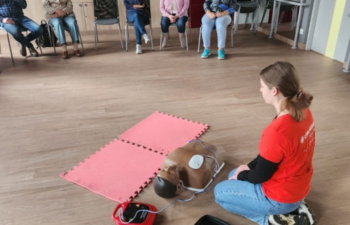 Wnętrze głównej sali w Centrum Aktywności Seniora. Na podłodze klęczy ratowniczka, która demonstruje seniorom użycie sprzętu do reanimacji.