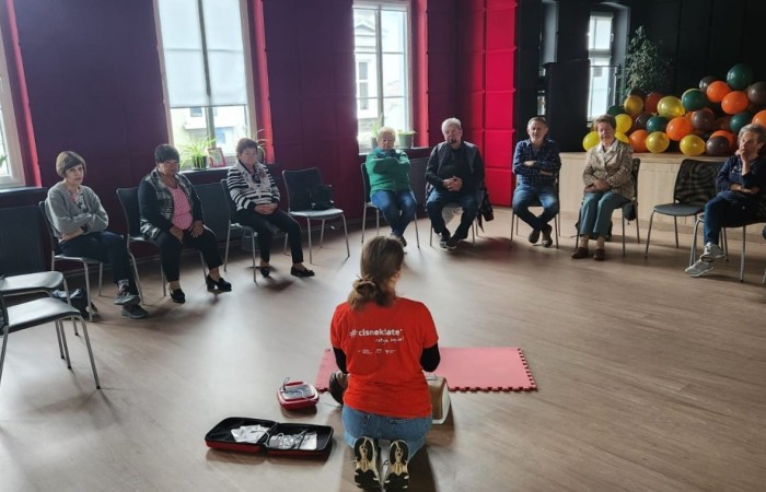 Wnętrze głównej sali w Centrum Aktywności Seniora. Na podłodze klęczy ratowniczka, która demonstruje seniorom użycie sprzętu do reanimacji. Przed nią na krzesłach w półokręgu siedzą seniorzy. W tle widać część sceny z balonami i wysokie okna Centrum.