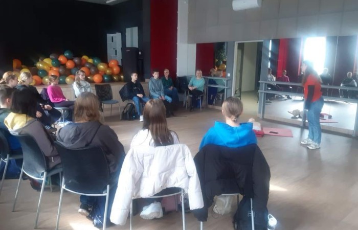 Kilkanaścioro uczniów siedzi na krzesłach w półokręgu i przysłuchuje się ratowniczce, która przekazuje wiedzę dotyczącą udzielania pierwszej pomocy. W tle od lewej widać scenę z balonami, otwarte drzwi do sali i ścianę luster do tańca i ćwiczeń.