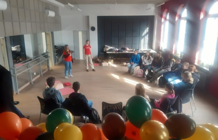 Widok na całą salę. Zdjęcie wykonano ze sceny. Na pierwszym planie widać balony. Za nimi na parkiecie w półokręgu na krzesłach siedzą uczniowie, a przed nimi stoją dwie ratowniczki. Przez wysokie okna od prawej strony do sali wpada światło słoneczne.