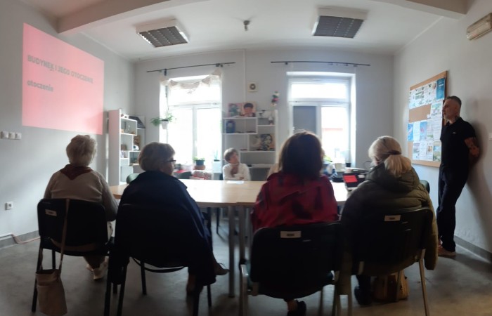 Warsztat dla seniorów pod tytułem Mój dom moja twierdza czy mój wróg. Zdjęcie przedstawia seniorów siedzących po obu stronach stołu i trenera prowadzącego spotkanie, stojącego i opartego o ścianę po prawej stronie zdjęcia. W tle widać dwa jasne okna.