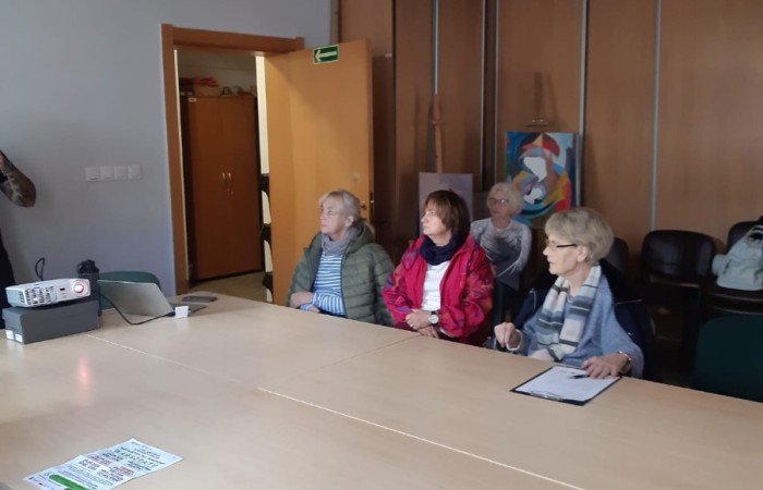 Warsztat dla seniorów Mój dom - moja twierdza czy mój wróg. Dużą część zdjęcia od dołu zajmują cztery jasne stoły złączone ze sobą. Przy nich siedzą trzy seniorki, a jedna seniorka siedzi z tyłu na krzesłach. Kobiety słuchają instruktora. 