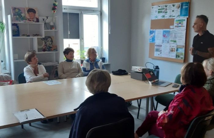 Warsztat dla seniorów Mój dom - moja twierdza czy mój wróg.  Wnętrze sali. Po obu stronach stołu siedzą seniorki, które słuchają instruktora prowadzącego spotkanie. W tle od lewej widać regał, jasne okno i tablicę korkową wiszącą na białej ścianie.