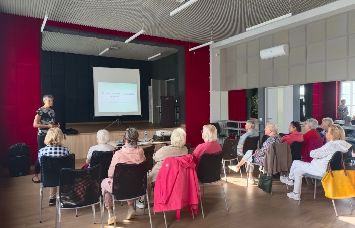 Zdjęcie przedstawia uczestników warsztatu pod tytułem Profilaktyka odleżyn, którzy słuchają wykładu pielęgniarki i oglądają kolejny slajd prezentacji tematycznej. Seniorzy siedzą na krzesłach przed sceną w głównej sali Centrum Aktywności Seniora.